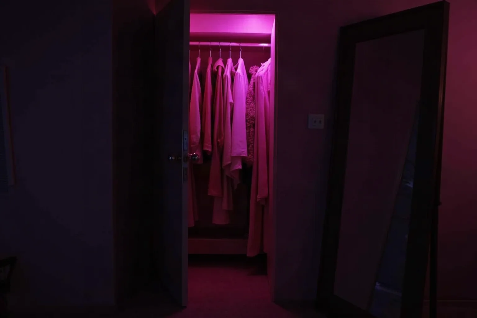An open wardrobe in a dim bedroom with silk garments and a mirror catching pink light